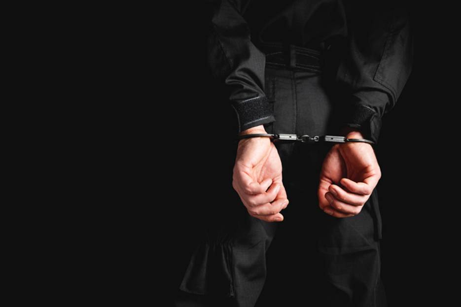 A person is wearing handcuffs behind their back. The image is set against a dark background, with the person dressed in black.